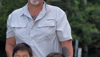 A bearded man wearing a light button-up shirt standing with two young children outdoors.