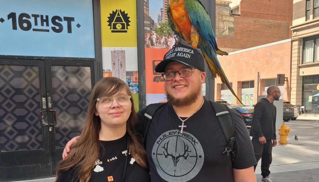 A smiling couple standing outside, with a vibrant blue and orange macaw parrot perched on top of the man's cap.