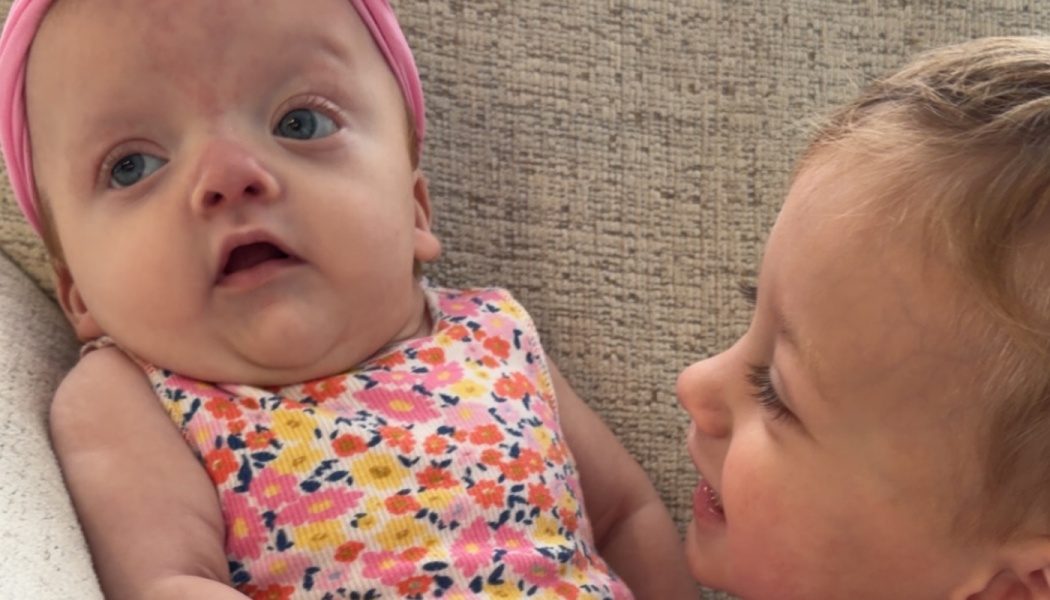 A smiling toddler looks closely at a baby wearing a pink headband and a colorful floral top.