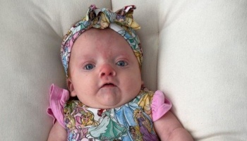 A baby with blue eyes wearing a colorful Disney Princess headband and matching outfit, lying on a cushion.