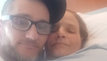 A man wearing glasses and a cap poses close to a woman resting on a pillow.