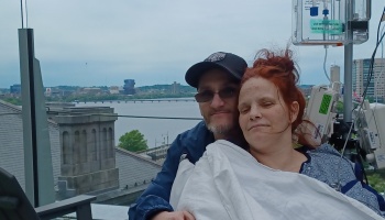 A smiling man embraces a red-haired woman wrapped in a blanket on a hospital terrace next to IV equipment.