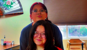 Two young women wearing glasses pose closely together, one smiling and the other making a playful pout.