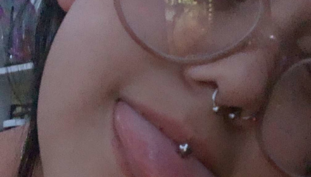 Close-up of a person with round glasses sticking out their tongue to show a silver tongue piercing and nose rings.