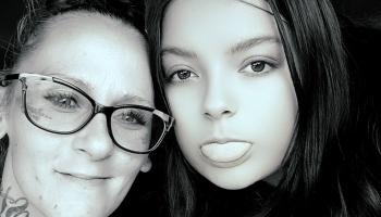 Black-and-white close-up of a smiling woman with glasses next to a young girl playfully sticking her tongue out.