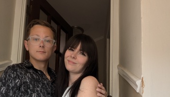 A man with clear glasses and a smiling woman with long dark hair and bangs posing together indoors.