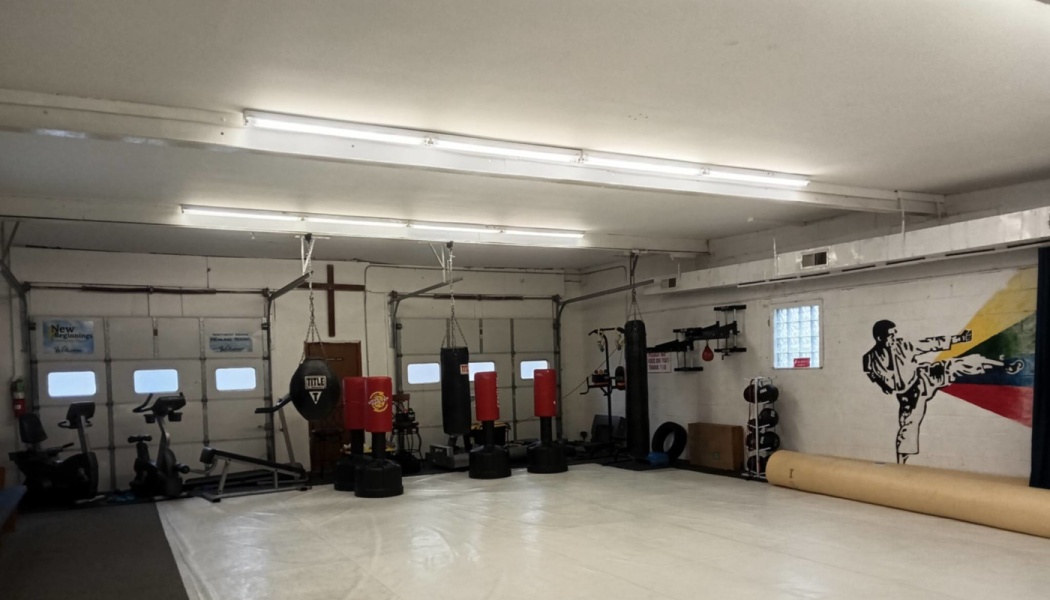 An indoor martial arts gym with white floor mats, hanging heavy bags, standing punching bags, and a wall mural.