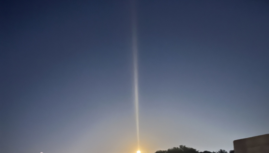 A bright vertical light pillar extending straight up into a dark blue sky from a glowing horizon.