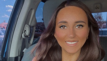 A smiling young woman with long brown hair and brown eyes sitting inside a car.