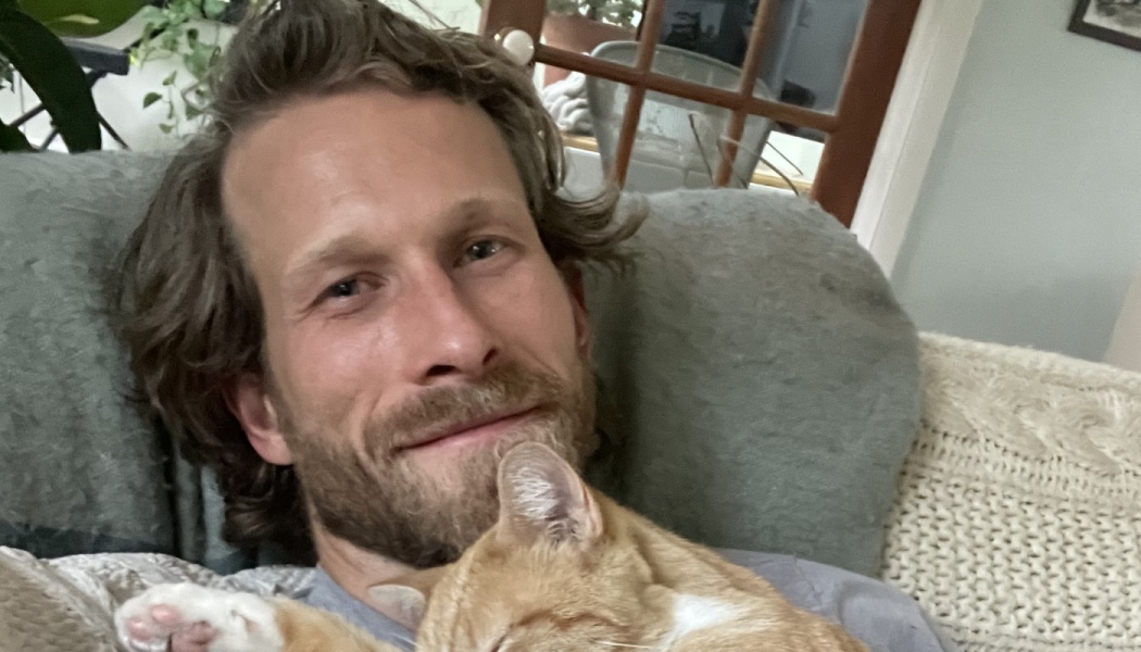 A smiling man lying down on a couch while an orange cat sleeps peacefully on his chest.