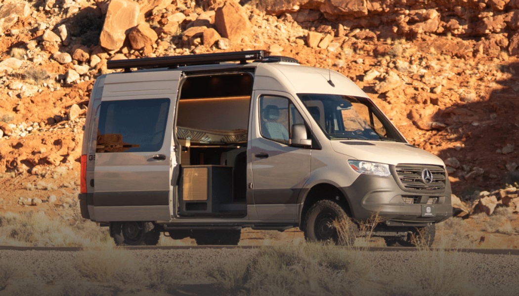 Grey camper van with an open side door revealing a cozy interior, parked against a red rock desert landscape.