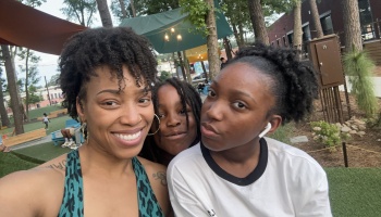 A smiling woman and two young girls posing for a selfie outdoors at a park with trees.