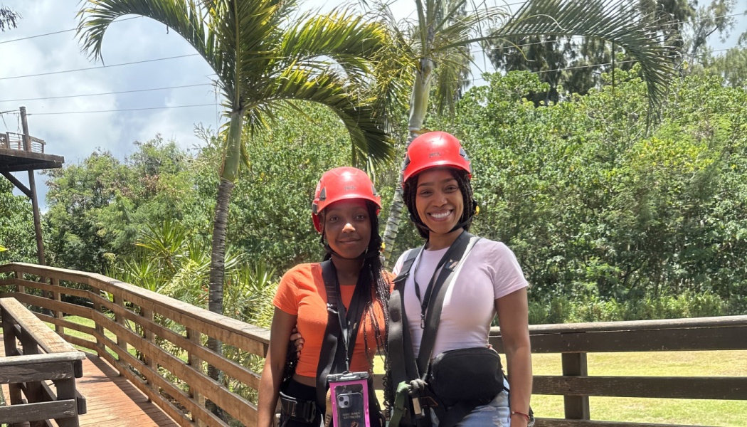Two smiling young women wearing red safety helmets and harnesses pose outdoors in a tropical setting.