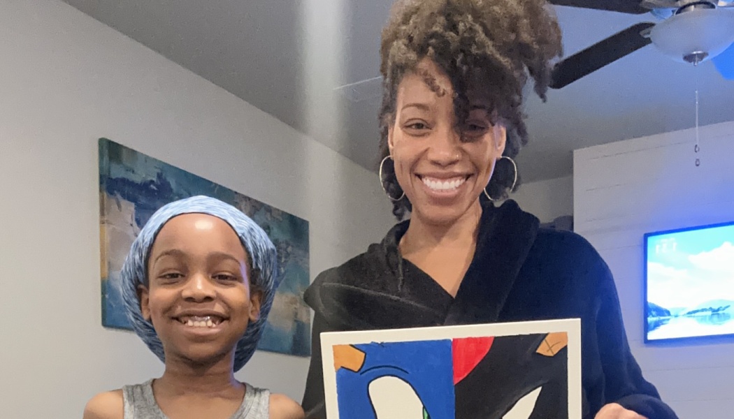 A smiling woman with curly hair and a young child hold up a colorful canvas painting indoors.