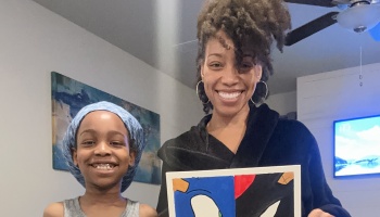 A smiling woman with curly hair and a young child hold up a colorful canvas painting indoors.