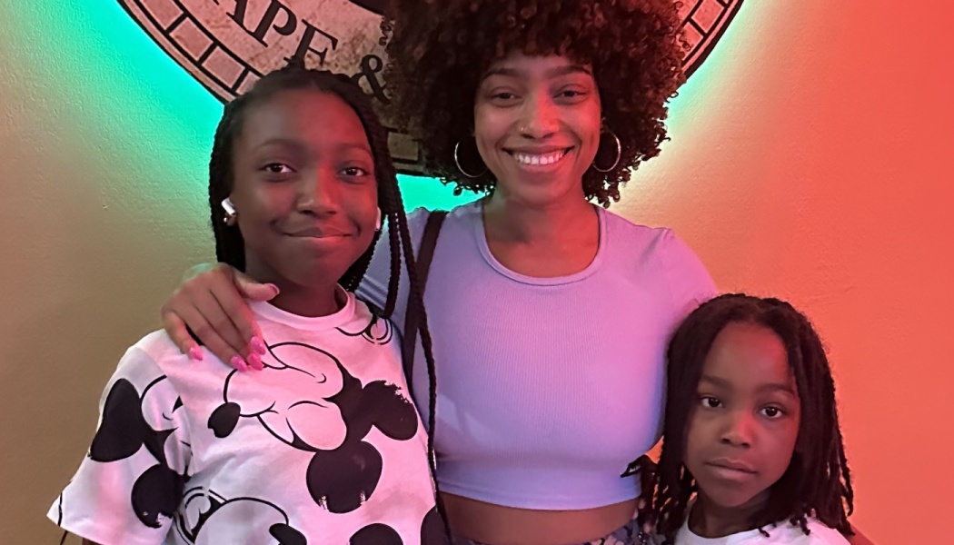 A smiling woman with curly hair poses happily with her two children under vibrant, colorful lighting.
