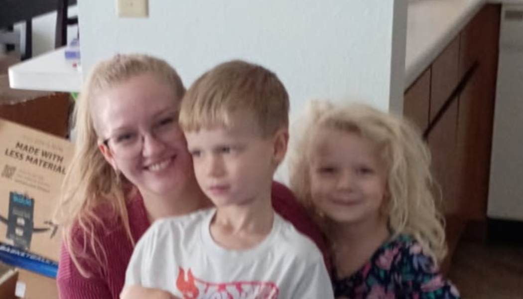 A smiling blonde woman with glasses posing happily with two young children indoors.