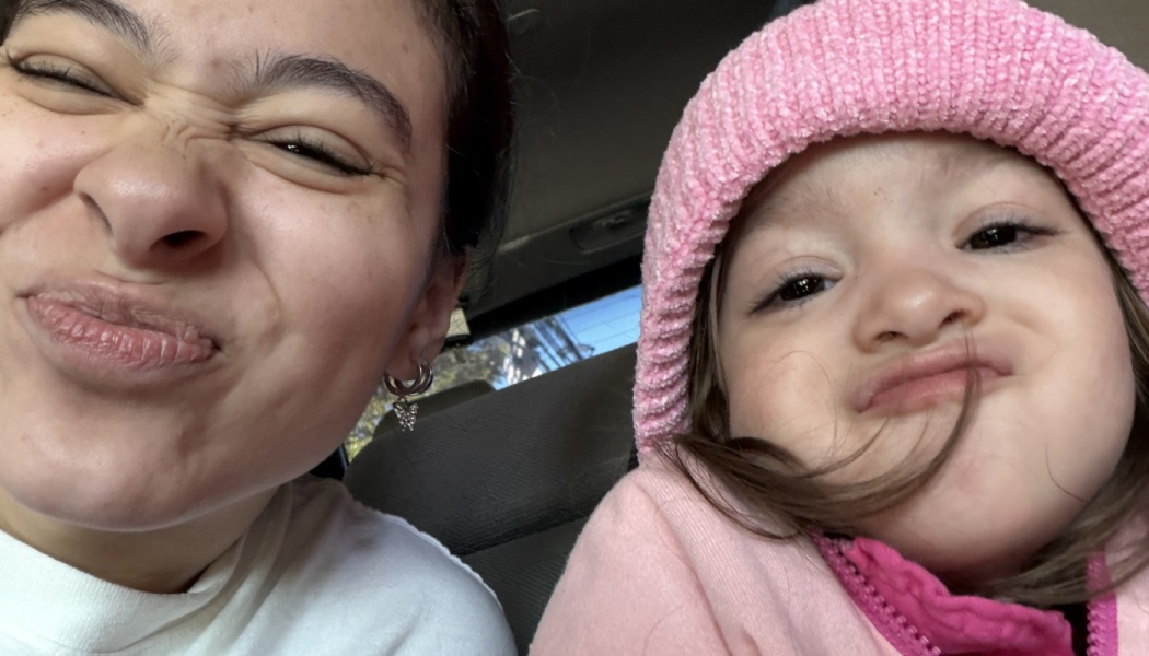 Close-up selfie of a young woman and a toddler girl in a pink beanie making funny scrunched faces in a car.