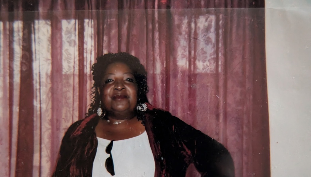 A smiling Black woman with dark curly hair wearing a white top and dark jacket in front of maroon curtains.