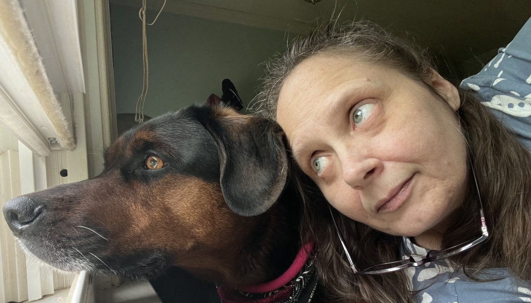 A woman rests her head against her black and tan dog as both look out a window.