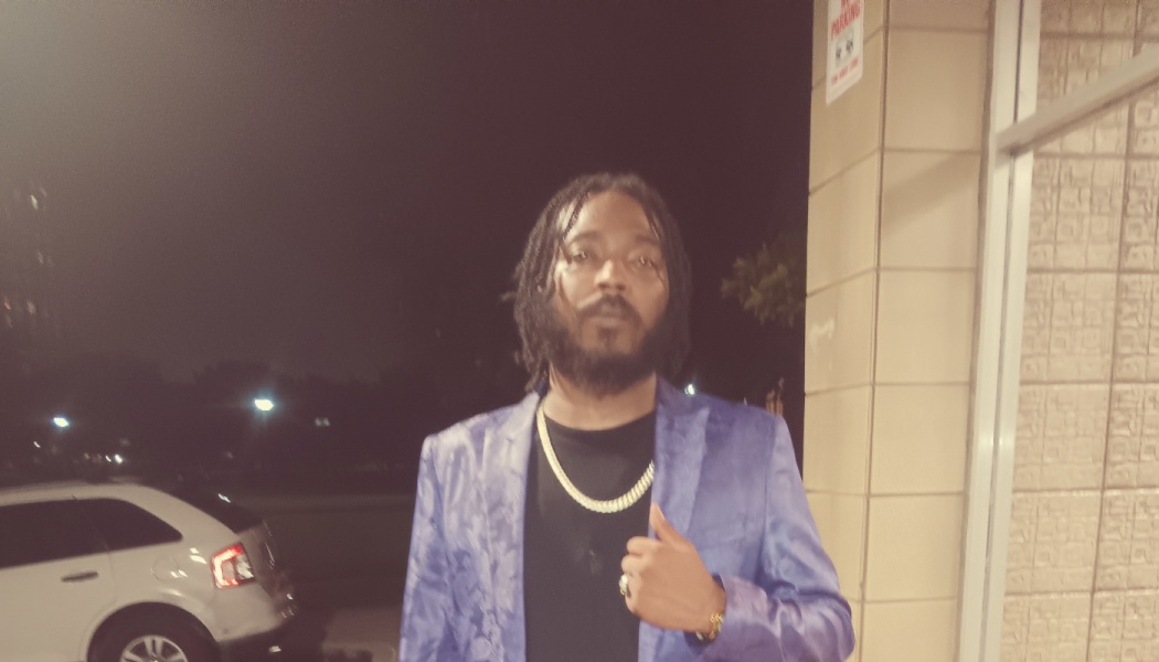 Man with dreadlocks and a beard wearing a patterned purple suit jacket and gold chain outdoors at night.