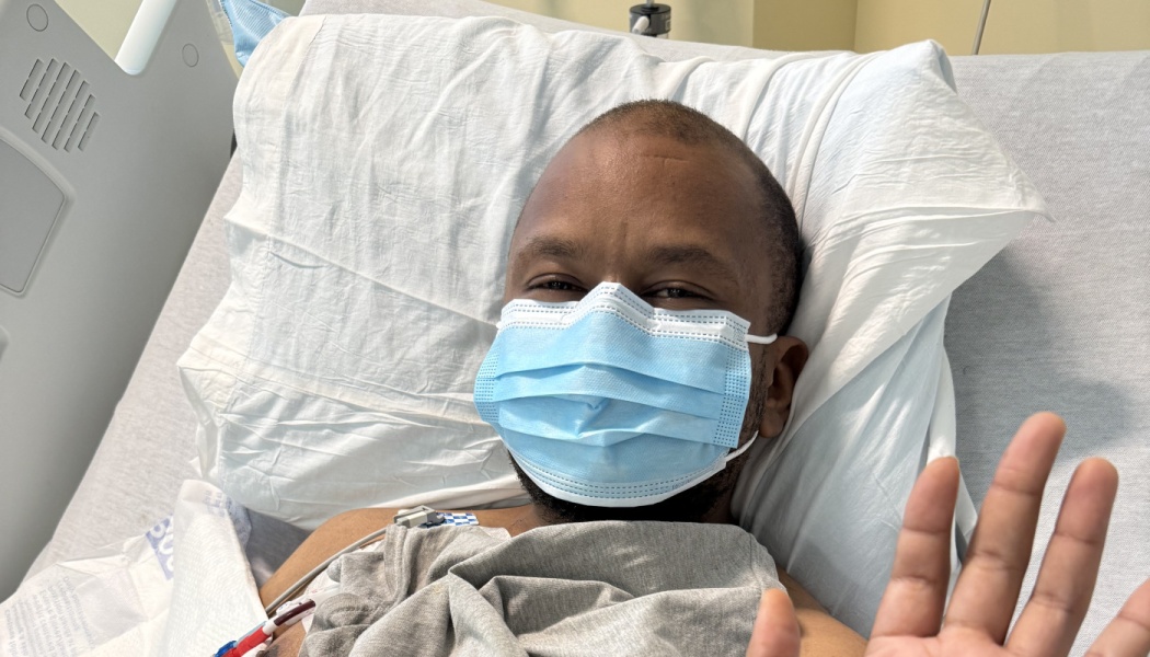 A man wearing a blue medical mask lies in a hospital bed and waves at the camera.