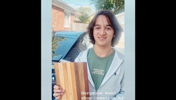 A smiling young man holding a handcrafted, multi-toned striped wooden cutting board outdoors.