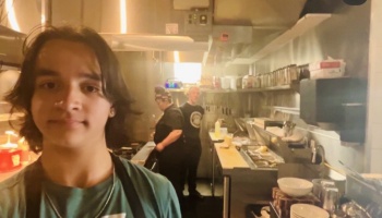 Young restaurant worker taking a selfie inside a commercial kitchen with colleagues working in the background.