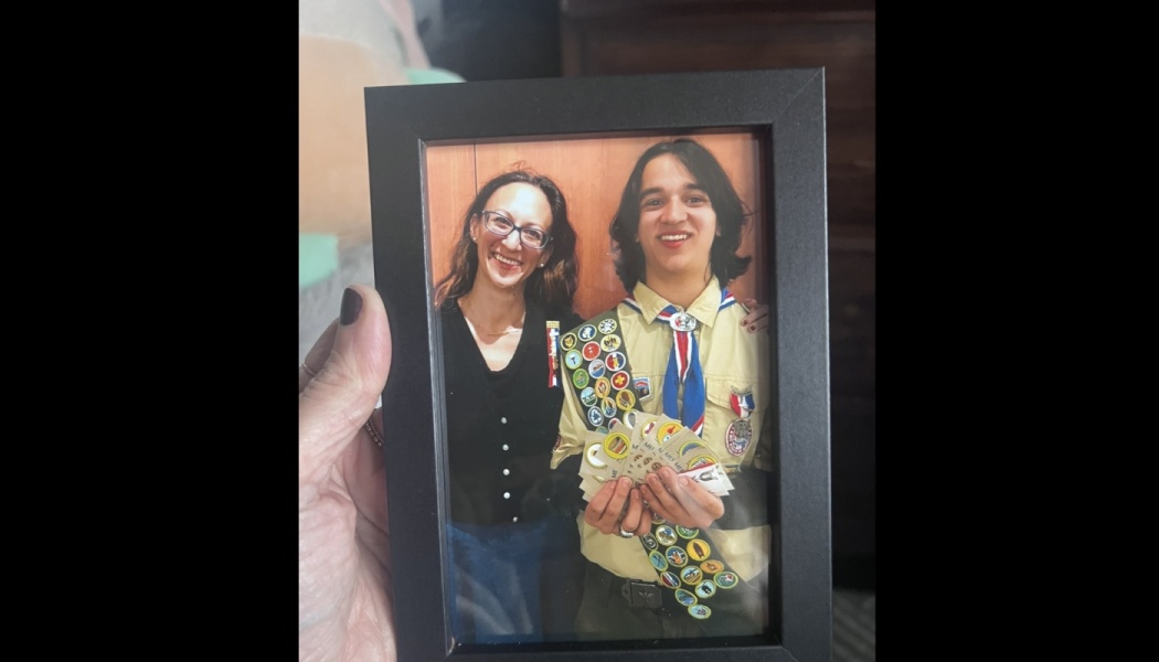 A hand holds a framed photo of a smiling woman standing next to a young scout displaying his merit badges.