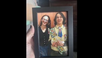 A hand holds a framed photo of a smiling woman standing next to a young scout displaying his merit badges.