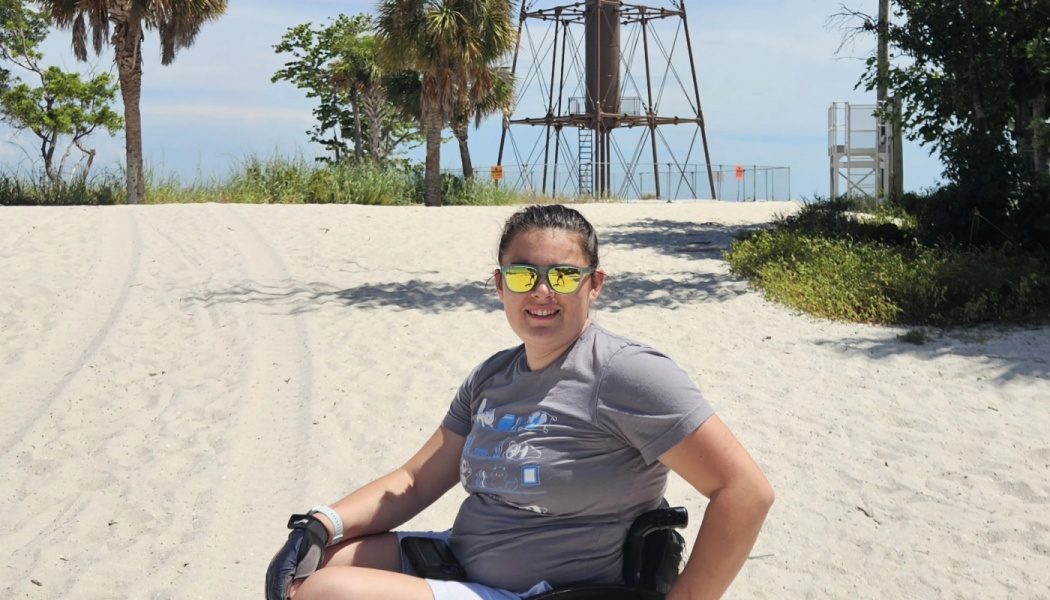 Smiling woman in sunglasses sitting in a wheelchair on a sandy beach with palm trees and a tower behind her.