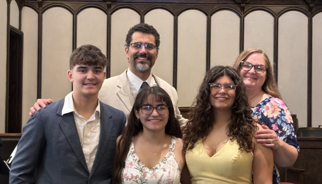 A smiling family of five, including parents and three young adult children, standing closely together indoors.