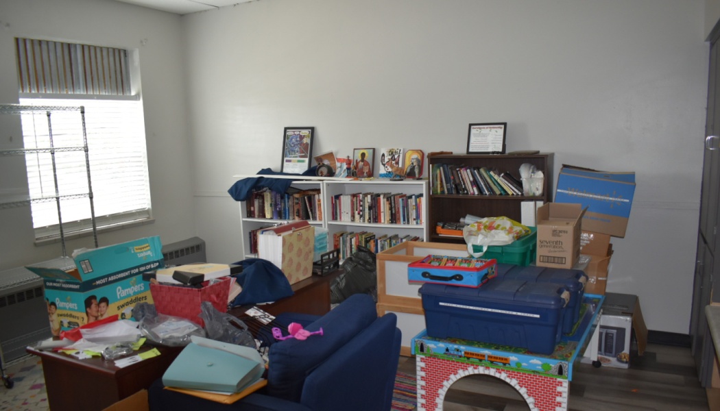 A cluttered room filled with packed bookshelves, cardboard boxes, plastic storage bins, toys, and furniture.