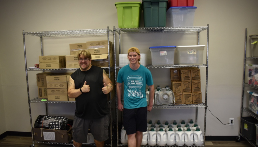 Two men stand in front of metal shelves stocked with cardboard boxes, plastic bins, and cleaning supplies.