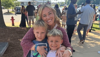 A smiling woman with blonde hair hugs her two young sons outdoors on a sunny day.