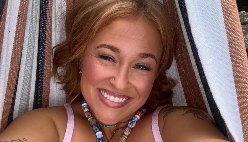 Smiling woman with short reddish hair lying in a striped hammock, wearing a colorful beaded necklace.