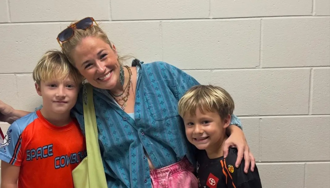 A smiling woman poses warmly between two young blonde boys against a white cinder block wall.