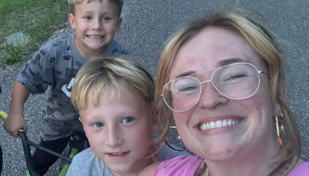 A smiling woman wearing glasses takes an outdoor selfie with two cheerful young boys.