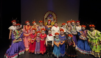 Performers in colorful Mexican folk dresses and Day of the Dead sugar skull face paint pose together on stage.