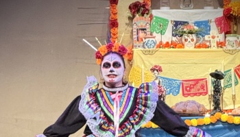 Performer in sugar skull makeup and colorful Mexican dress in front of a decorated Día de los Muertos altar.