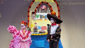 Two children in Day of the Dead costumes and skull face paint stand on stage in front of a decorated altar.
