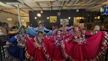 Five young dancers in vibrant, colorful traditional Mexican folklorico dresses spreading their wide ruffled skirts.