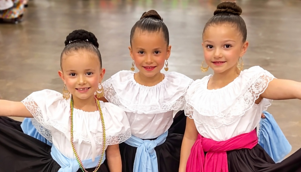 Three young girls smiling in traditional Folklorico dresses with white ruffled tops and hair in neat high buns.