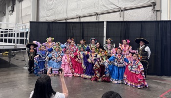 Children wearing colorful Mexican folklorico dresses pose together while a woman takes a phone photo.