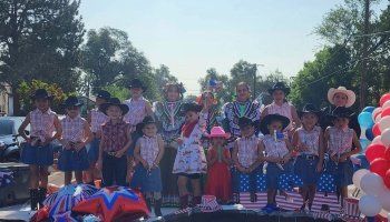 Children in cowboy hats and traditional dresses pose on a patriotic, USA-themed parade float.