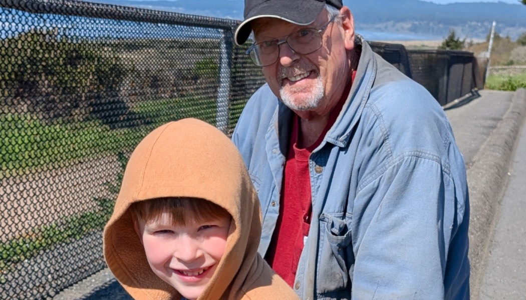 A smiling man in glasses and a black cap stands next to a young boy in an orange hoodie outdoors.