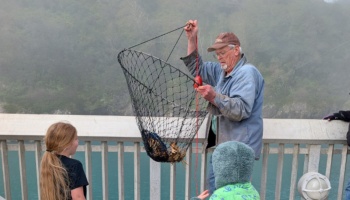 An older man shows a freshly caught crab in a large net ring to two children watching on a misty pier.