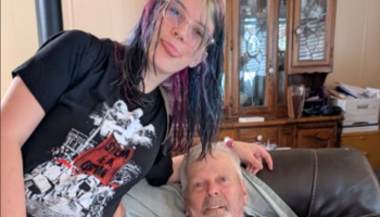 Young person with dyed hair and glasses leaning affectionately over an elderly man resting on a couch.