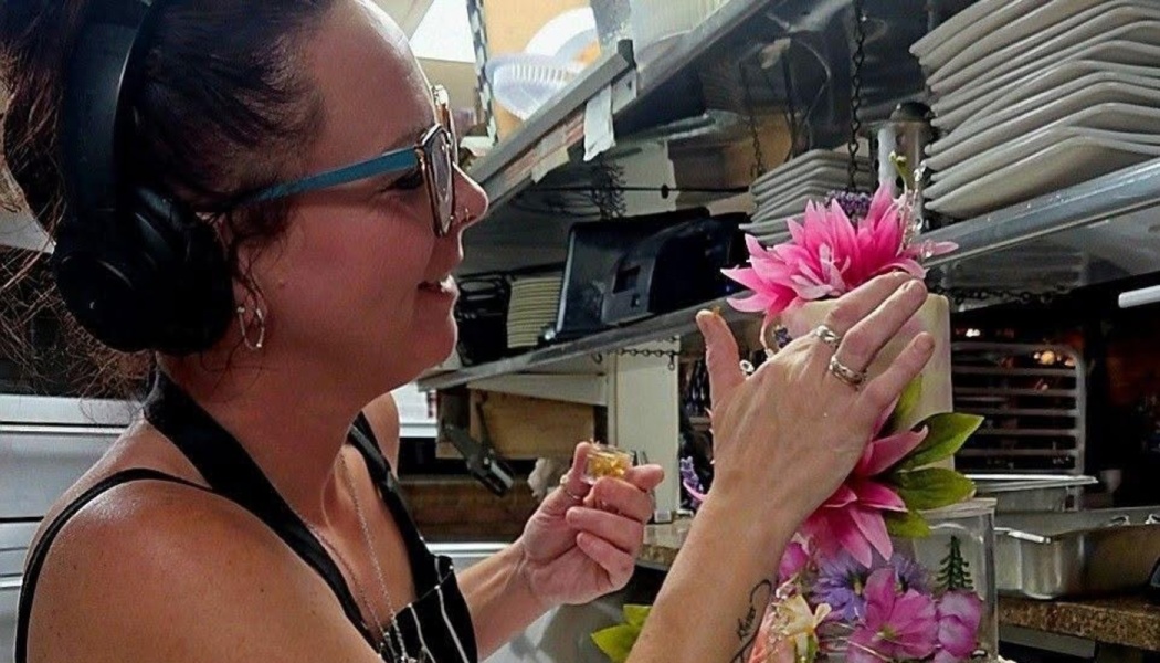 Woman wearing headphones and glasses carefully decorates a cake with bright pink flowers in a kitchen.