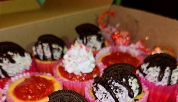 An open box filled with gourmet mini cheesecakes topped with Oreo cookies, whipped cream, and fruit jam.